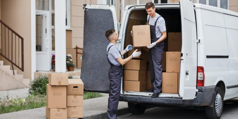 two-young-handsome-movers-wearing-uniforms-are-unloading-the-van-full-of-boxes-house-move-mover-1-e1694426635971